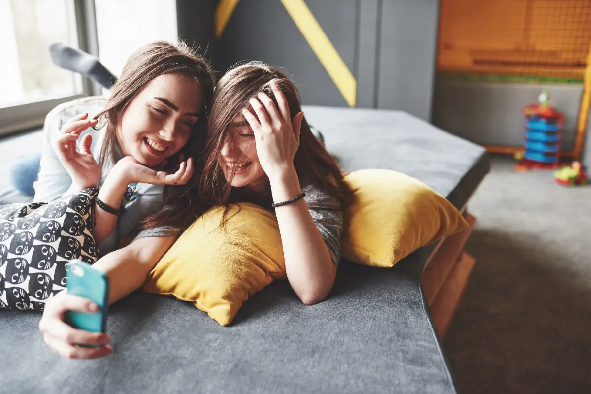 Kanepede sarı yastıklara uzanıp telefonla birlikte selfie çeken iki kadın ev arkadaşı